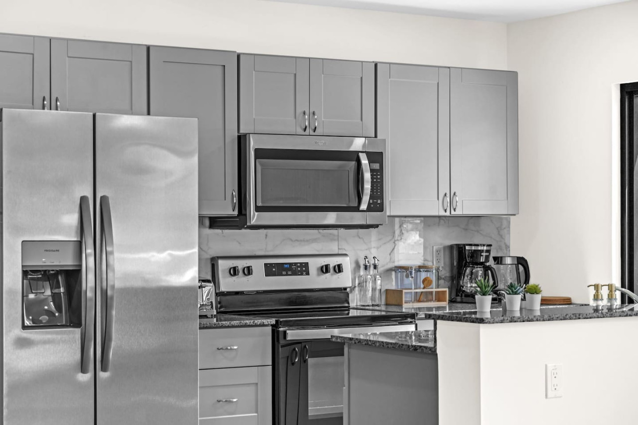 This modern kitchen features a spacious island with cabinetry, sleek white countertops, stainless steel appliances, open shelving, and stylish lighting.