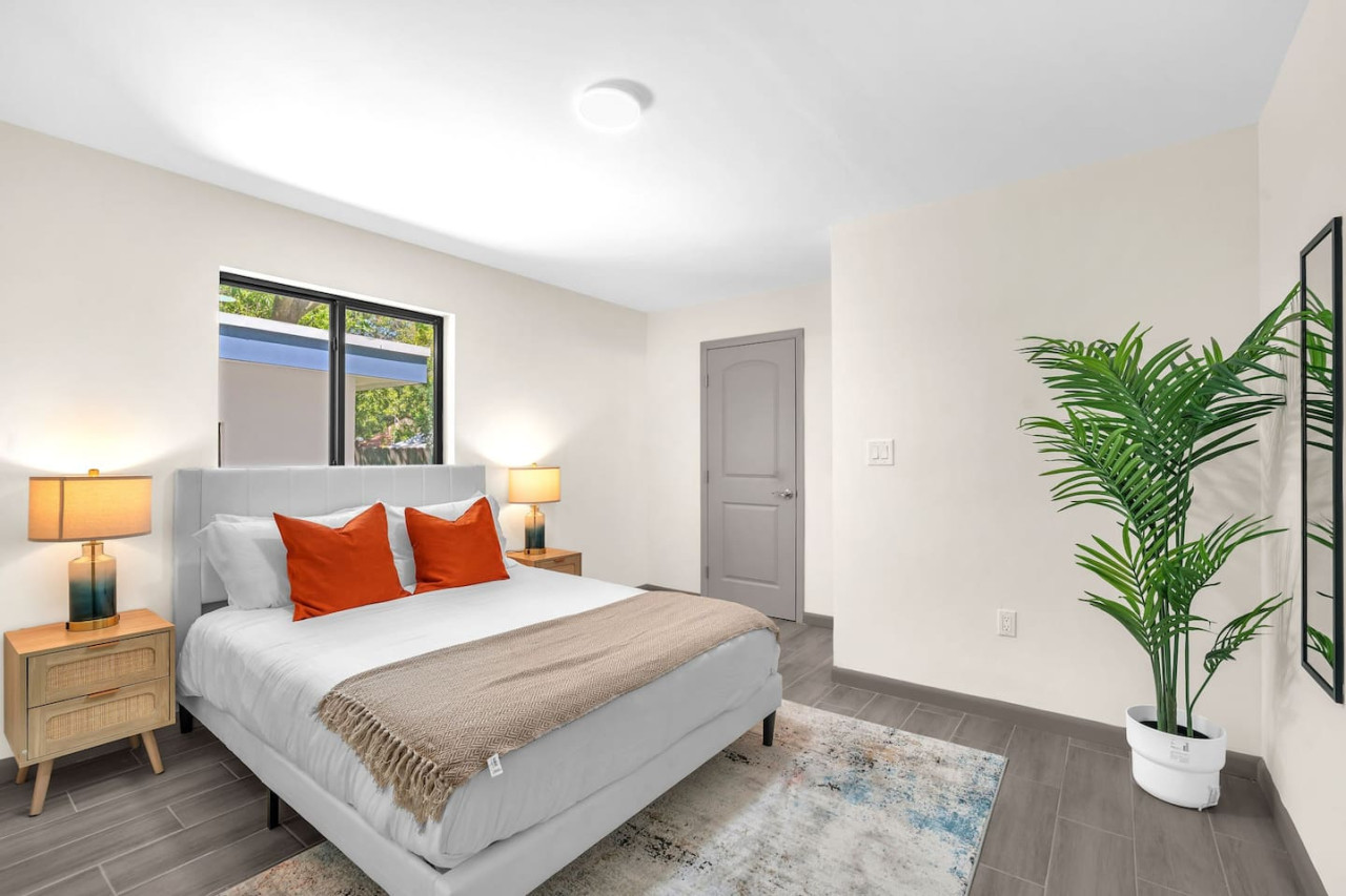A bright and inviting bedroom with a modern design, featuring crisp white bedding, vibrant orange accent pillows, and natural light streaming through the large windows.
