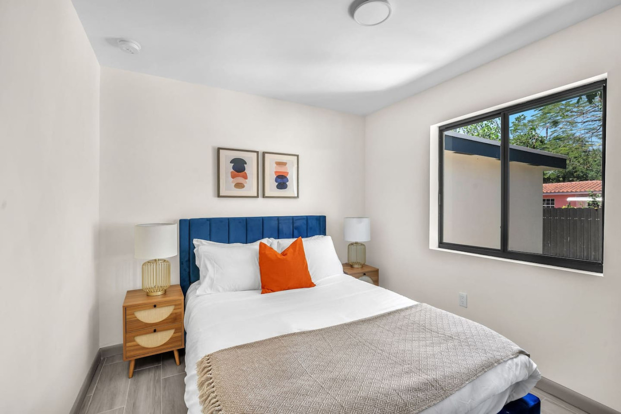 Bright bedroom featuring a soft, cushioned blue headboard, vibrant orange accent pillows, and natural lighting through a large window.