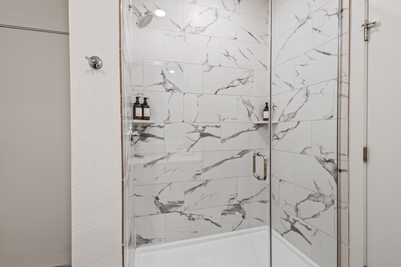 Refreshing modern shower featuring sleek glass door, elegant marble-patterned tiles, and convenient wall shelves for your essentials.