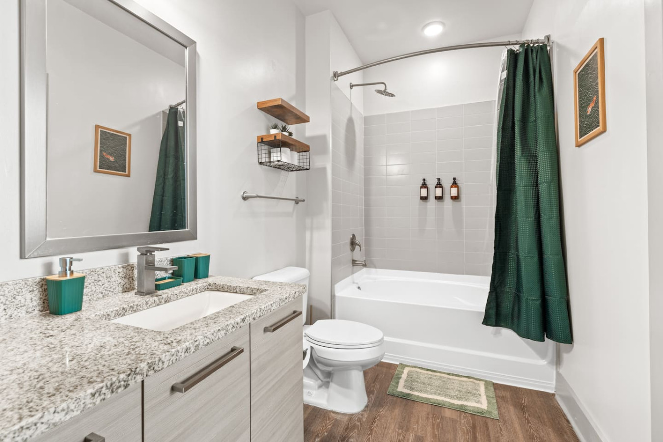 Clean, well-appointed bathroom with granite vanity, large mirror, and a shower-tub combo with rainfall showerhead. Complimentary shampoo, conditioner, and body wash provided. Floating wood shelves and matching teal accessories throughout.
