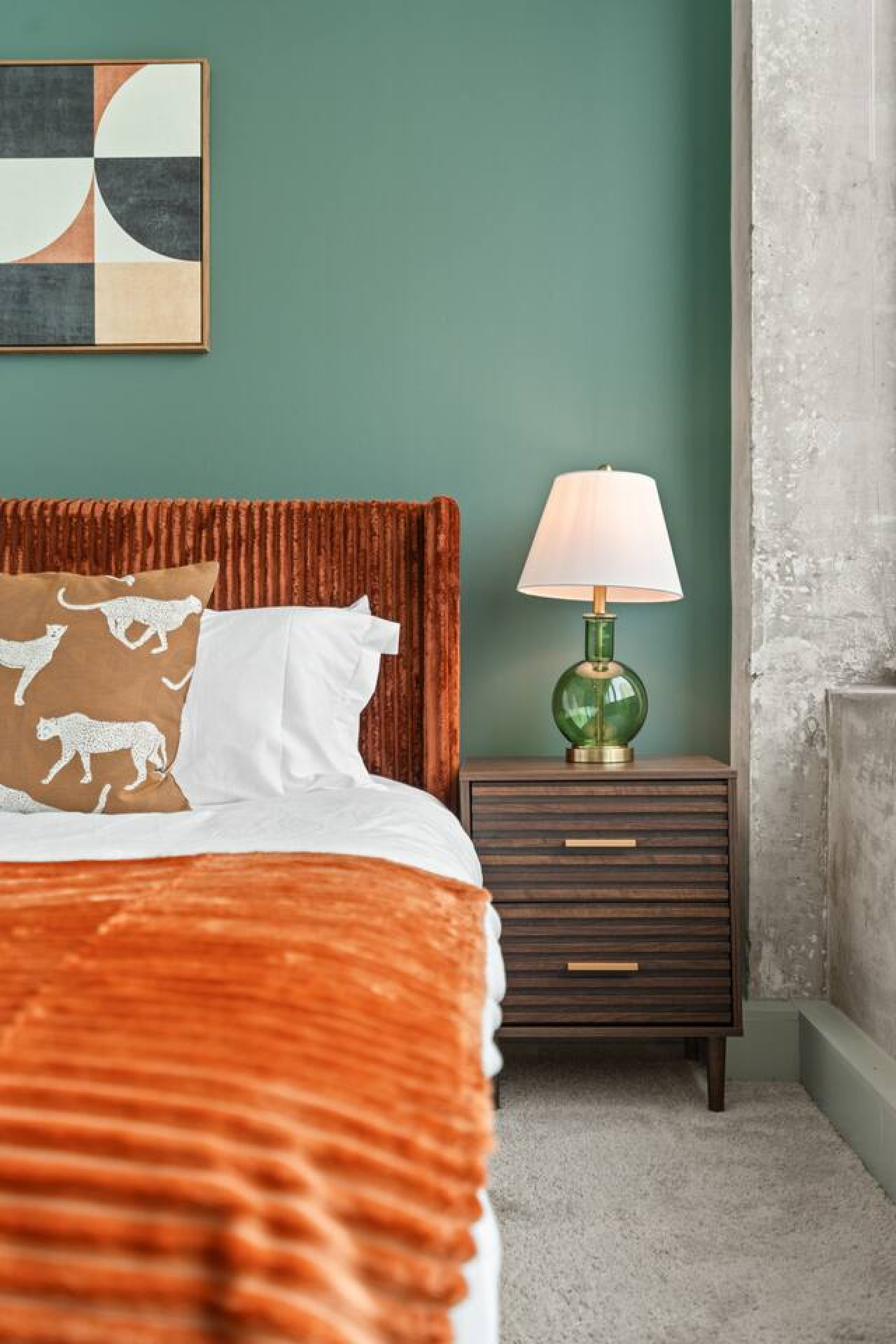 Bedroom details — rich corduroy headboard, playful leopard-print accent pillows, and a green glass lamp on a walnut nightstand. Exposed concrete column adds authentic loft charm to the cozy, design-forward space.