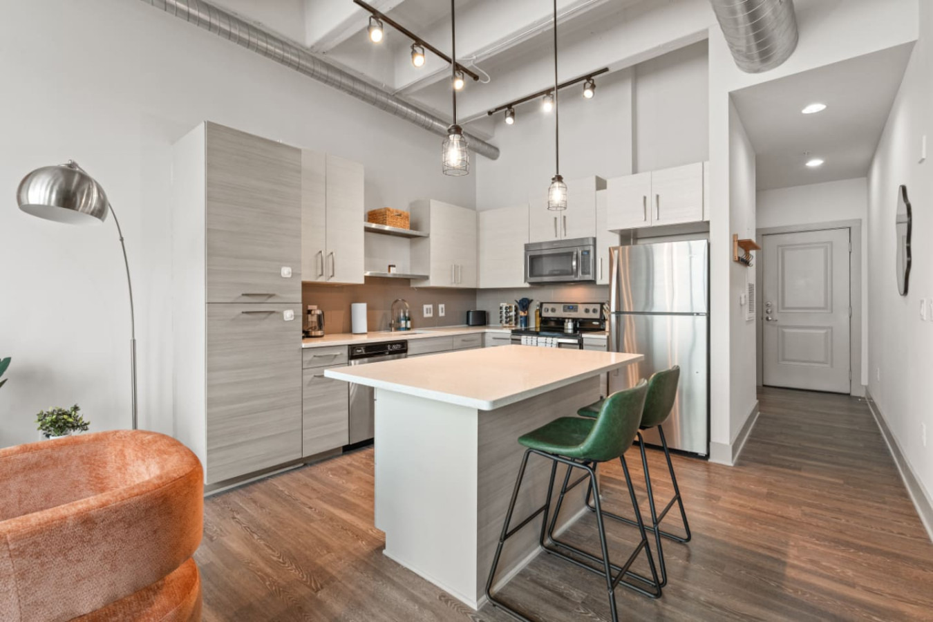 Wide view of the open kitchen flowing into the living area and entry hallway. Soaring ceilings with track and pendant lighting, a spacious island with green leather bar stools, and a clean modern layout throughout.
