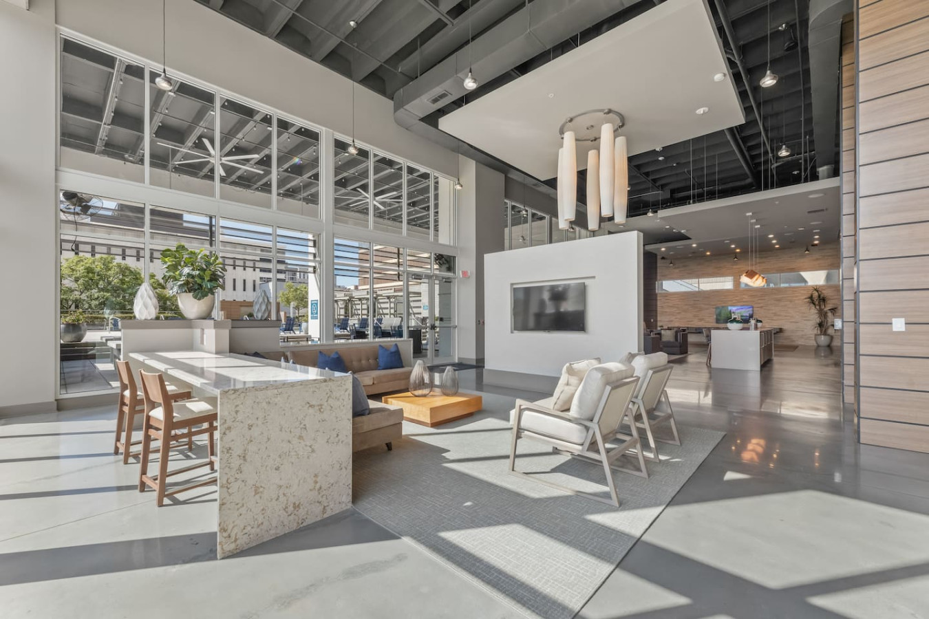 Grand resident lounge with soaring ceilings, designer pendant lighting, and a mix of seating and workspace areas. Features a large TV, communal table, and floor-to-ceiling windows overlooking the city