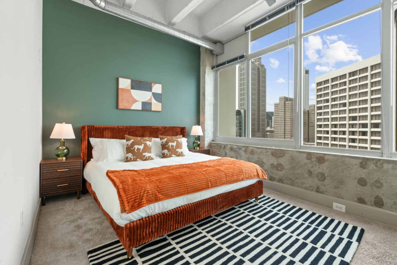 King-size bedroom with a plush corduroy upholstered bed, crisp white linens, and a terracotta throw. Wake up to stunning downtown Atlanta skyline views through oversized windows. Matching nightstands and modern art complete the space.