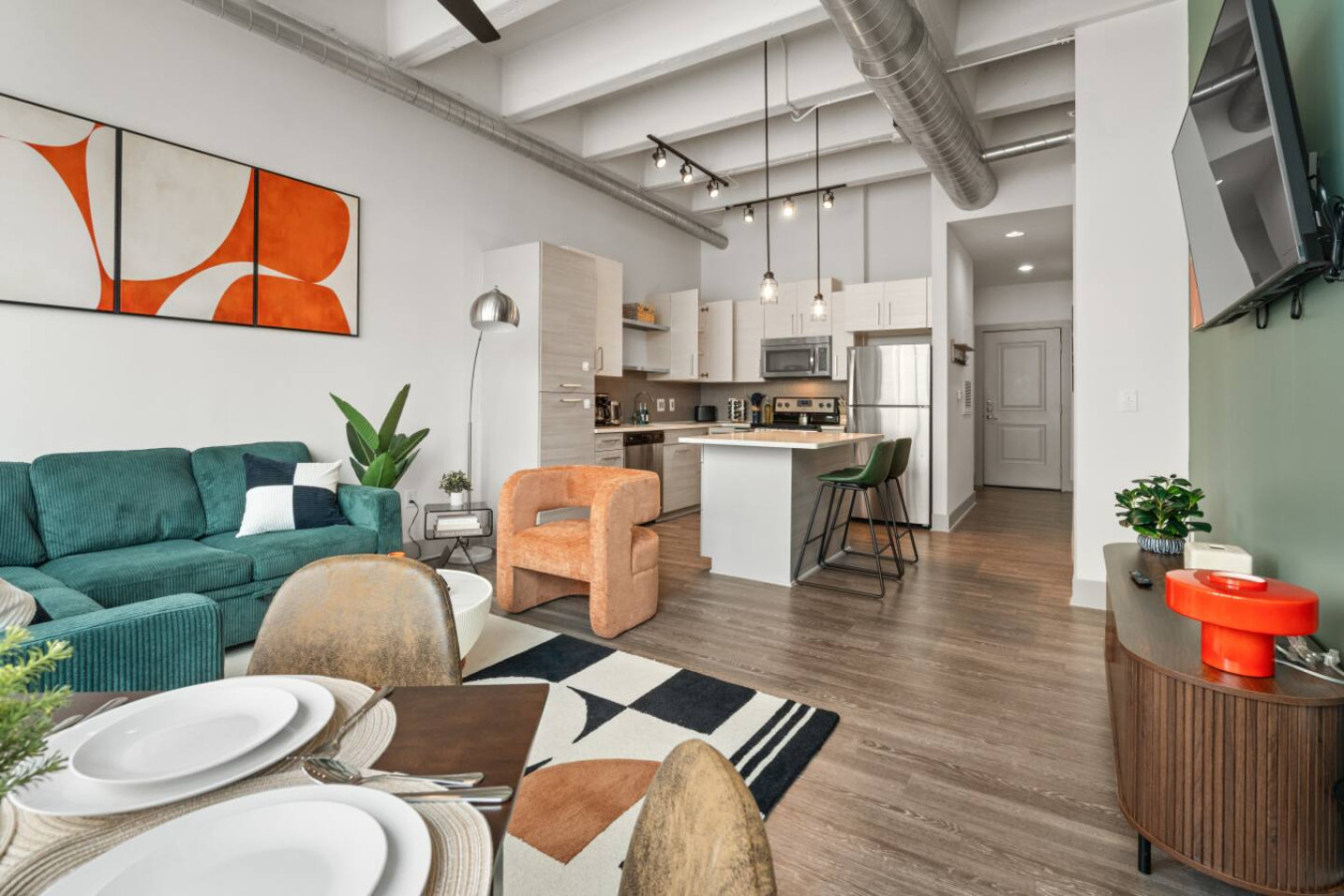 Open-concept living and kitchen area with high ceilings, exposed ductwork, and pendant lighting. Fully equipped kitchen with stainless steel appliances and breakfast bar. Dining table set for four, bold modern art, and designer furnishings throughout