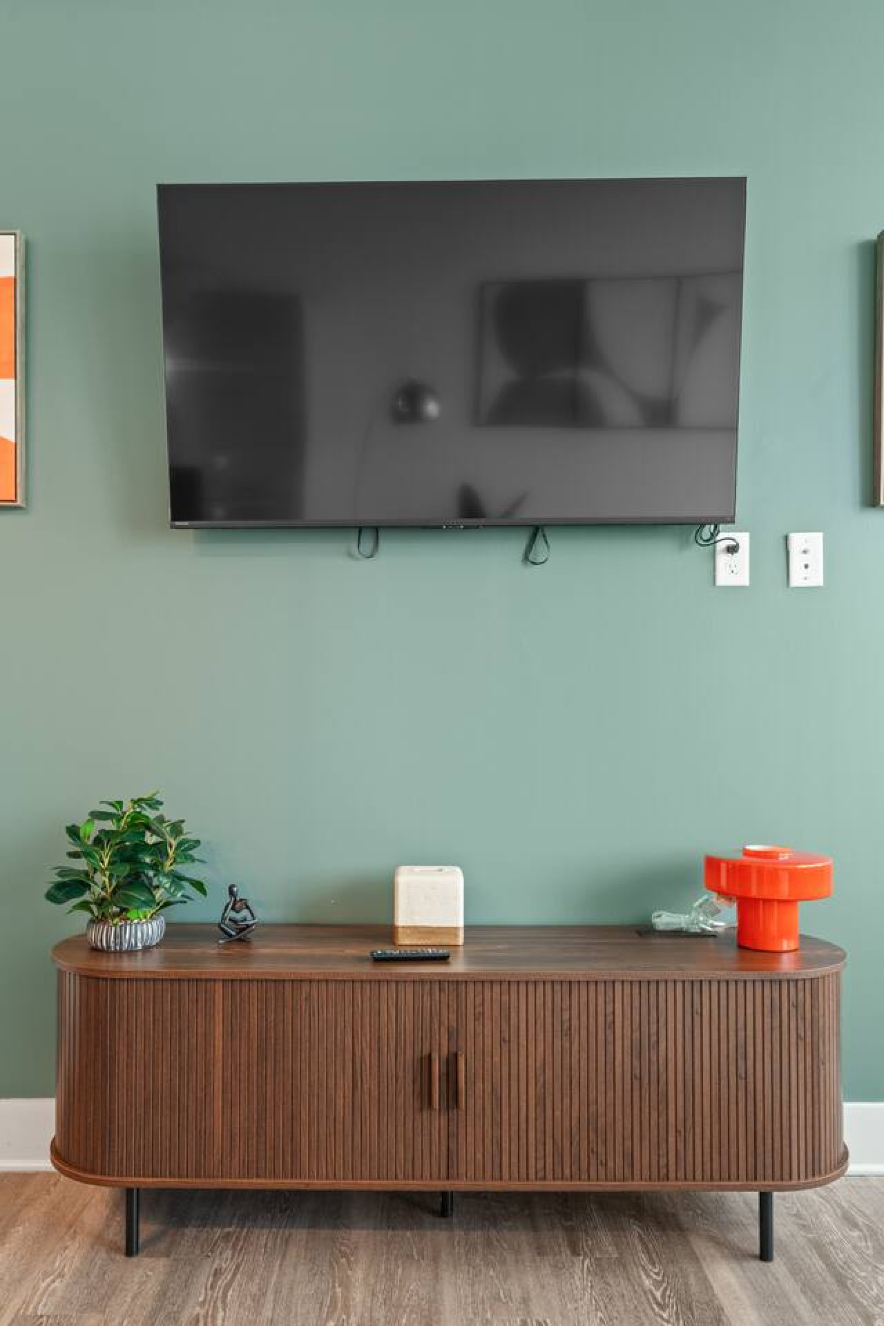 Wall-mounted 55" smart TV on a sage green accent wall with a fluted walnut media console below. Stream your favorites or catch the game after a day exploring Atlanta. Styled with greenery and modern décor accents.