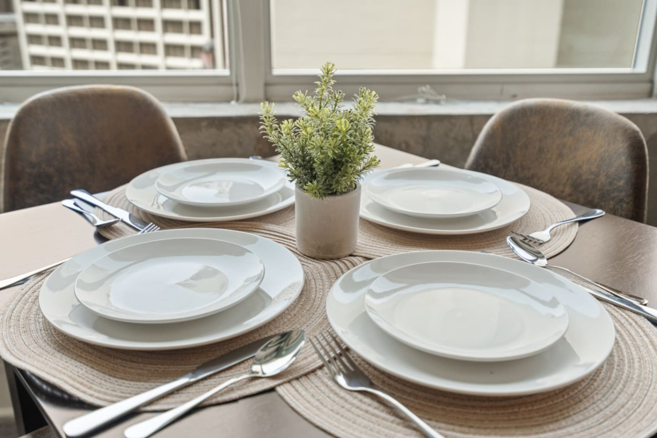 Dining table set for four with white dinnerware, woven placemats, and a fresh greenery centerpiece — all with downtown Atlanta views right outside the window. Perfect for shared meals with your group.
