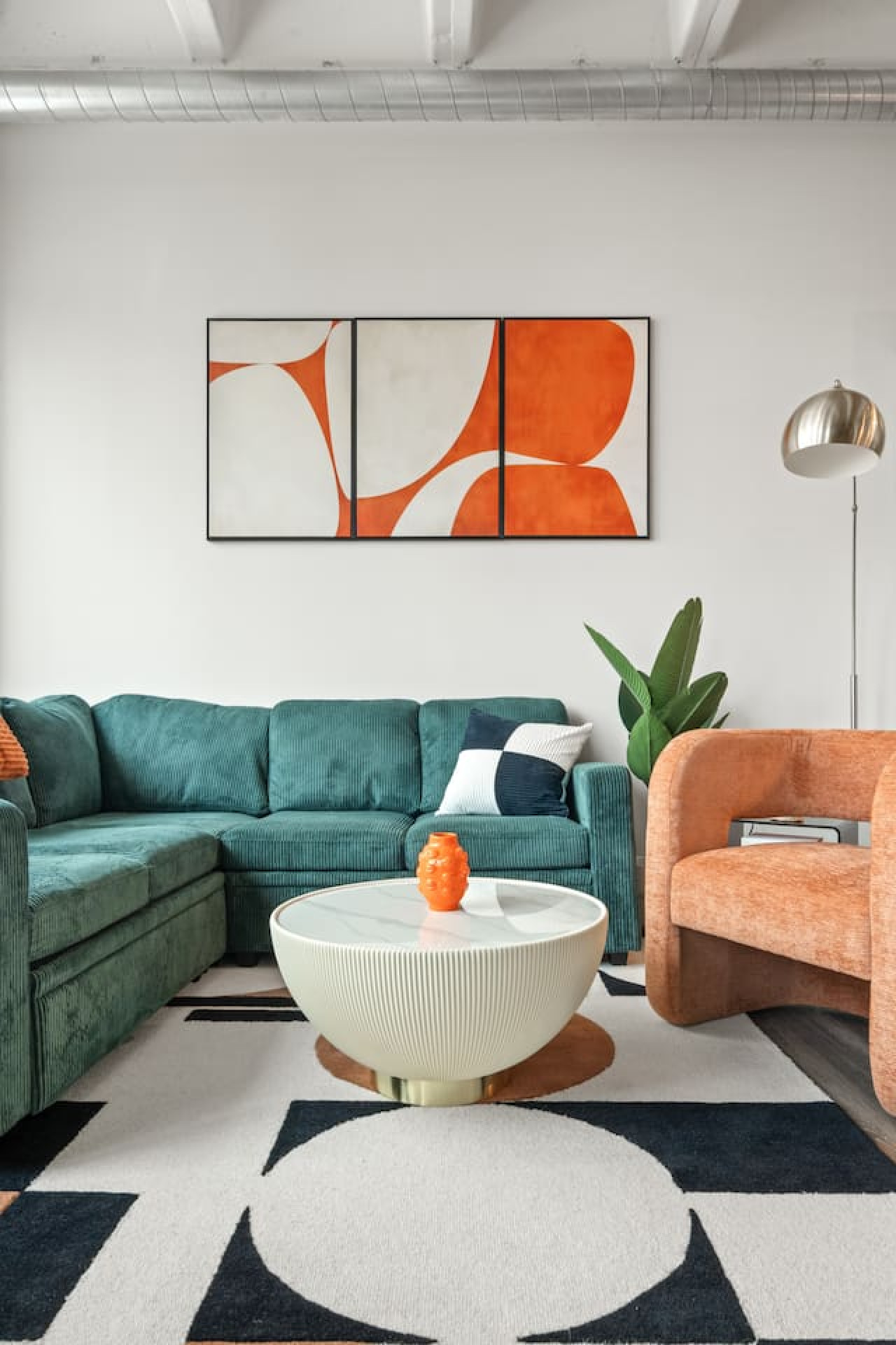 Curated seating area with a deep teal corduroy sectional, terracotta accent chair, and marble-top coffee table. Bold graphic artwork and a geometric area rug tie the mid-century modern palette together under loft-style exposed beams.