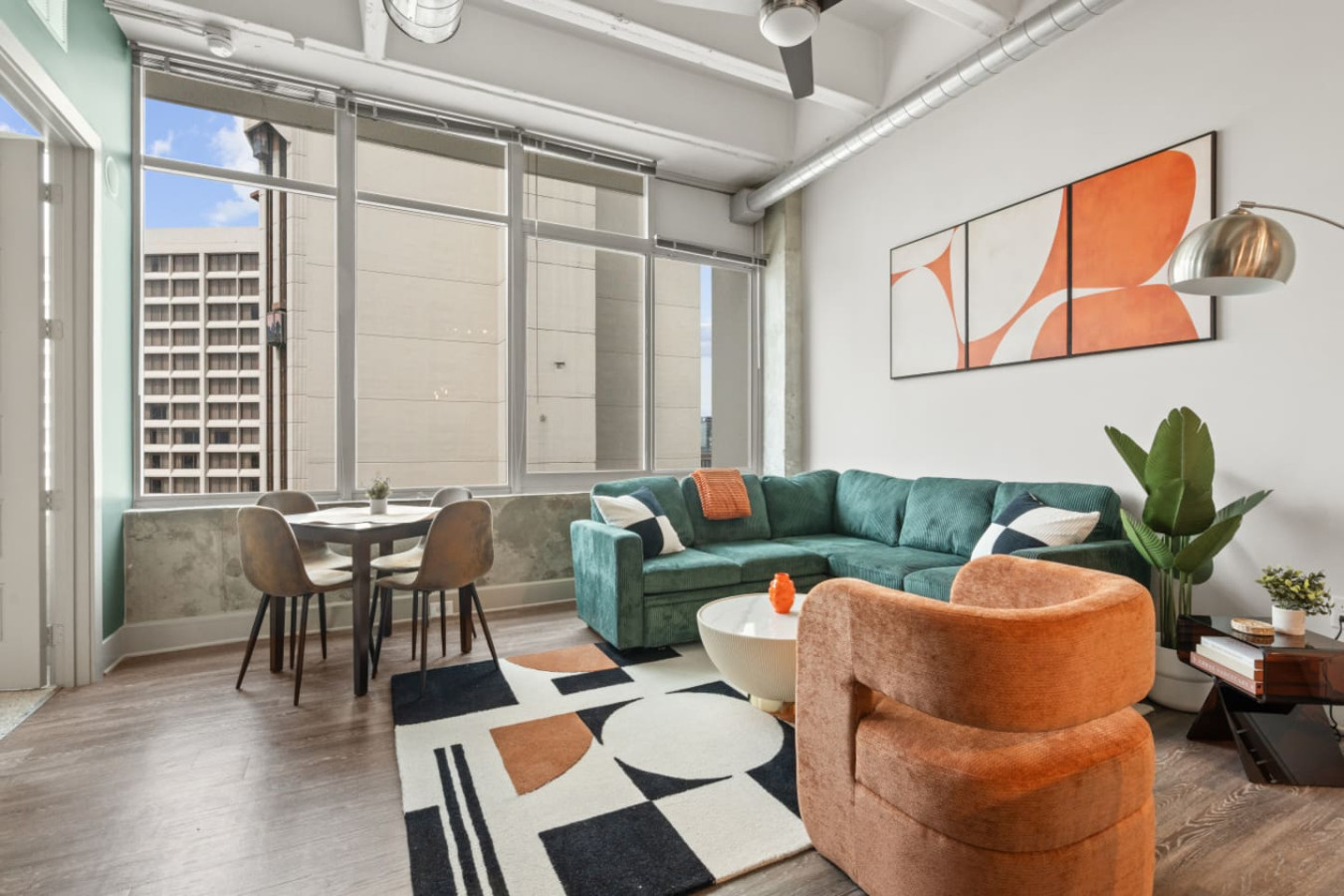 Sun-drenched living area with oversized windows framing downtown Atlanta high-rise views. A dining table for four sits beside the windows, perfect for morning coffee while taking in the city. Exposed concrete columns add authentic loft character.