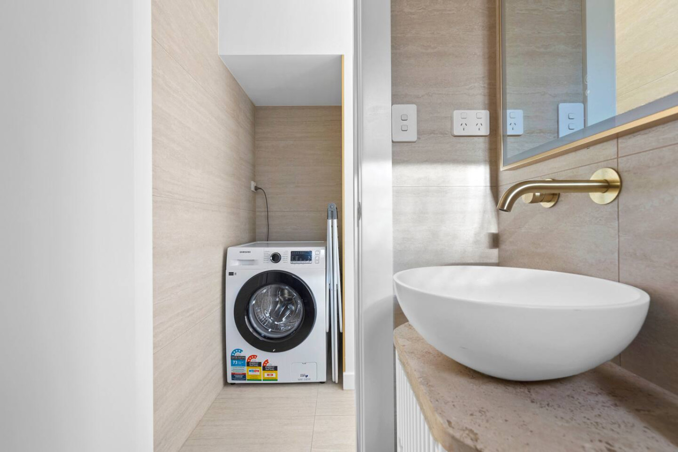 Compact yet stylish laundry nook integrated into the bathroom, featuring a sleek washer, gold accents, and a modern vessel sink. By Superhostly