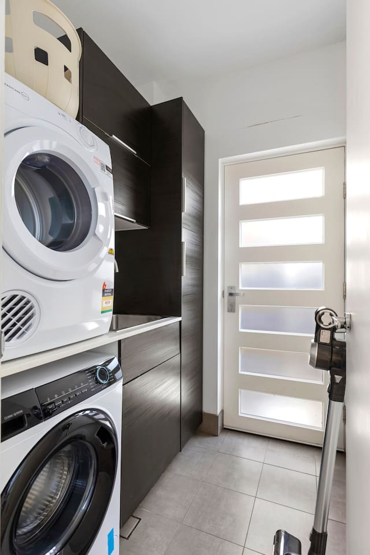 A sleek and functional laundry space featuring a stacked washer and dryer, ample dark cabinetry for storage, and a modern frosted glass door that brings in natural light while maintaining privacy.