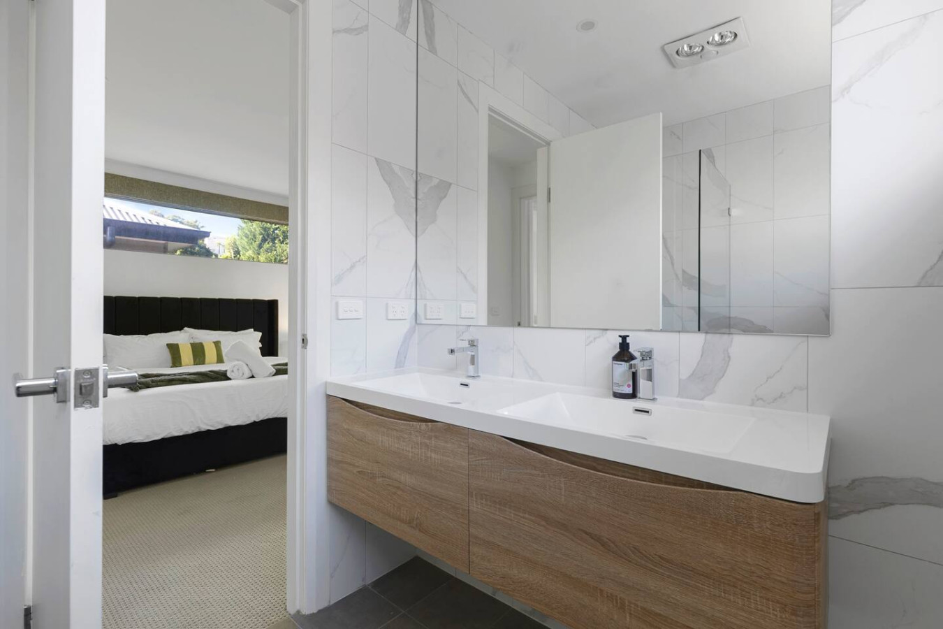 A sleek and modern bathroom featuring a stylish double vanity with wooden cabinetry, marble-tiled walls, and a large mirror, seamlessly connecting to a luxurious bedroom. By Superhostly