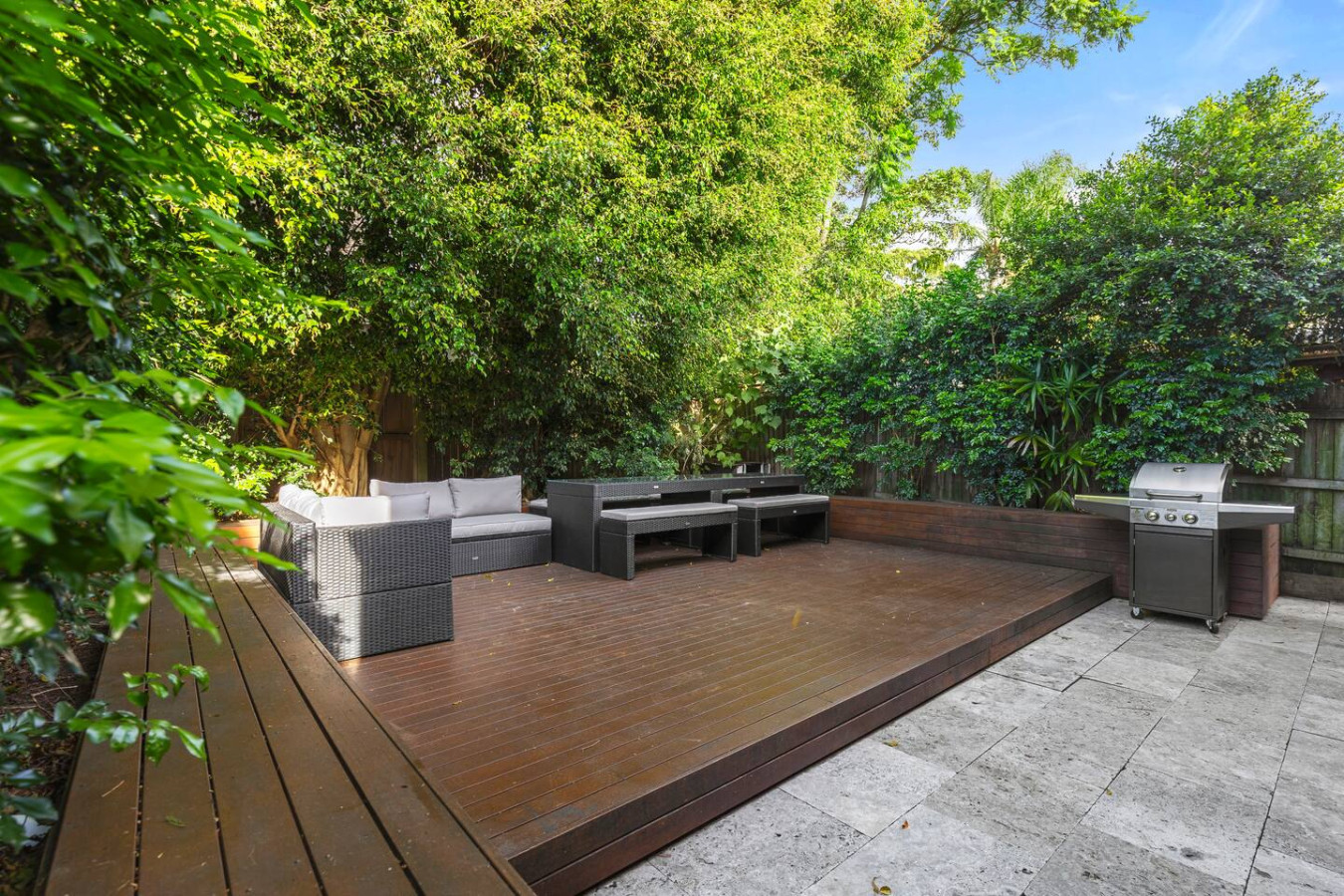A stunning outdoor entertainment space featuring a stylish wooden deck, modern rattan seating, and a sleek BBQ setup—all surrounded by lush greenery for a private retreat. By Superhostly