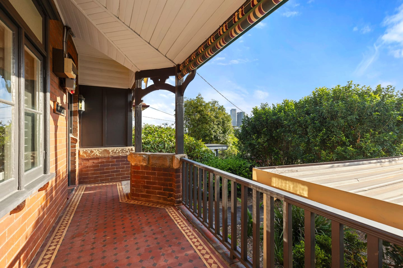 A charming covered veranda with intricate tiled flooring and brick accents, overlooking lush greenery with a glimpse of the city skyline in the distance—offering a perfect blend of heritage charm and urban convenience.