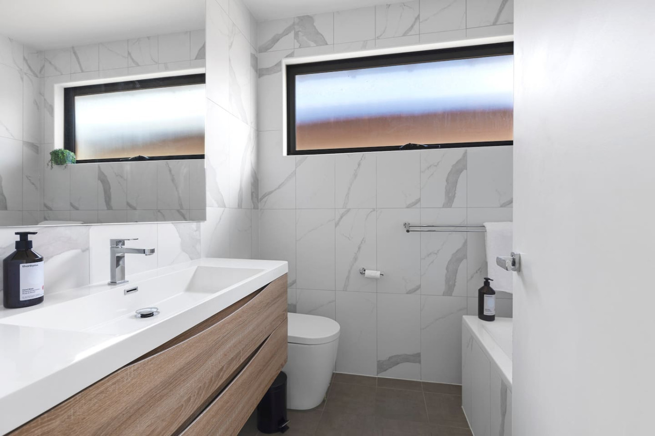 A contemporary bathroom featuring a stylish double vanity with a sleek wooden finish, marble-tiled walls, a modern toilet, and a bathtub, illuminated by frosted windows for natural light and privacy. By Superhostly
