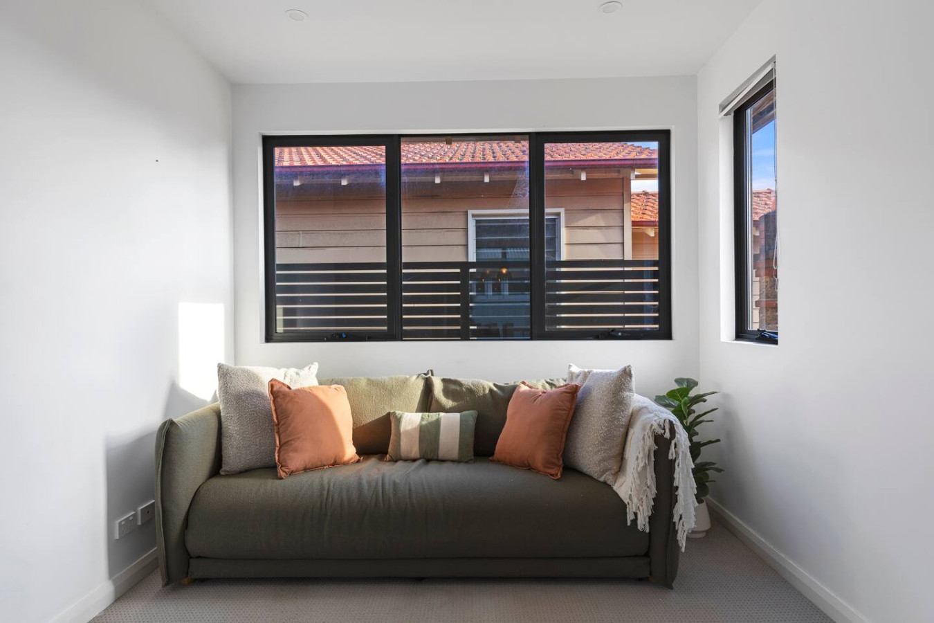A cozy sitting area featuring a stylish olive-green sofa adorned with warm-toned accent pillows and a soft throw, positioned by large windows that invite natural light and offer a view of neighboring rooftops.