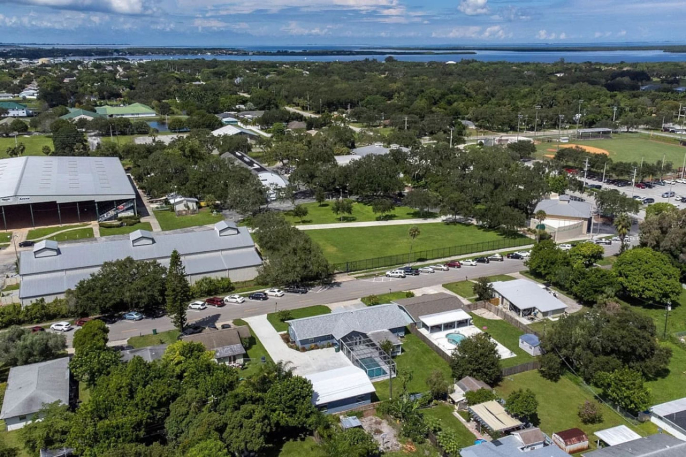 An aerial view from behind the house, with the fairgrounds' arena buildings and a ball field visible next door, and open water visible in the distance.