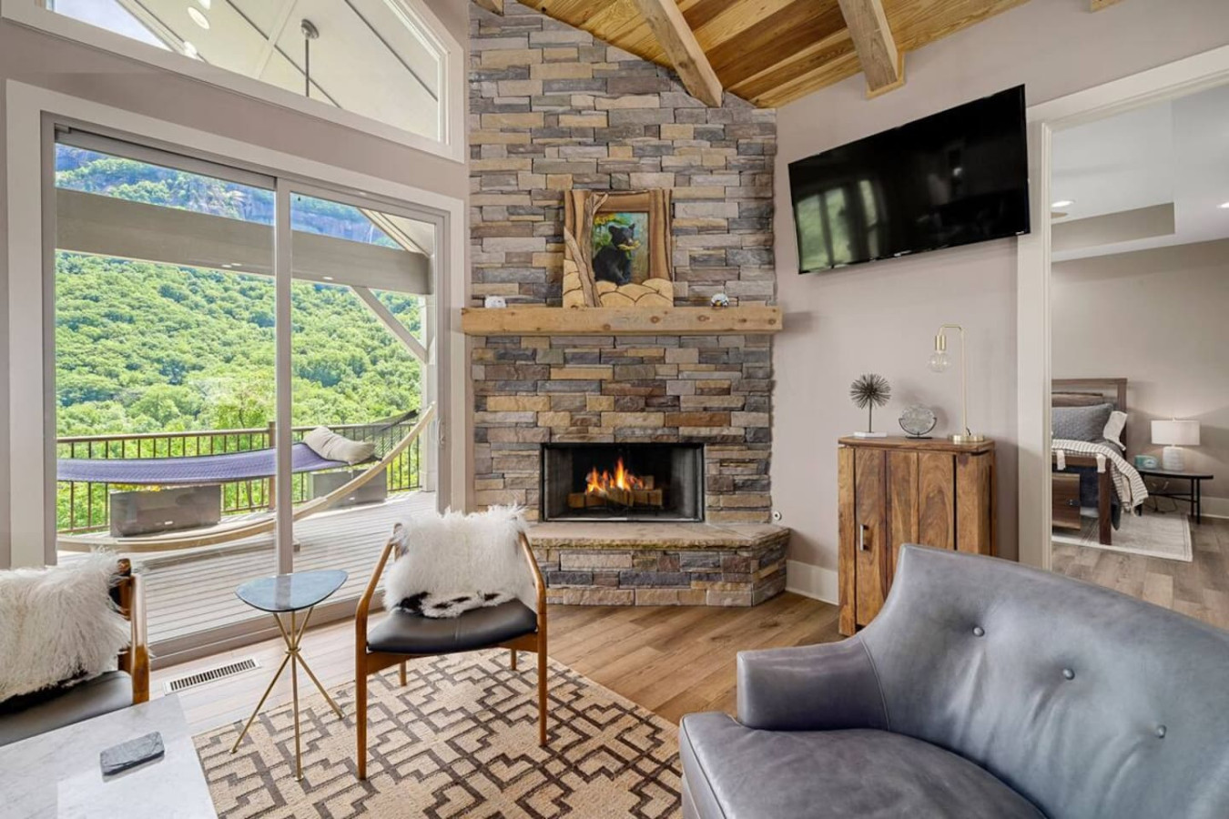 Cozy mountain living room with stone fireplace, vaulted wood ceilings, and plush seating. Large windows showcase stunning mountain views. Perfect for relaxing by the fire or enjoying nature's beauty from comfort indoors.