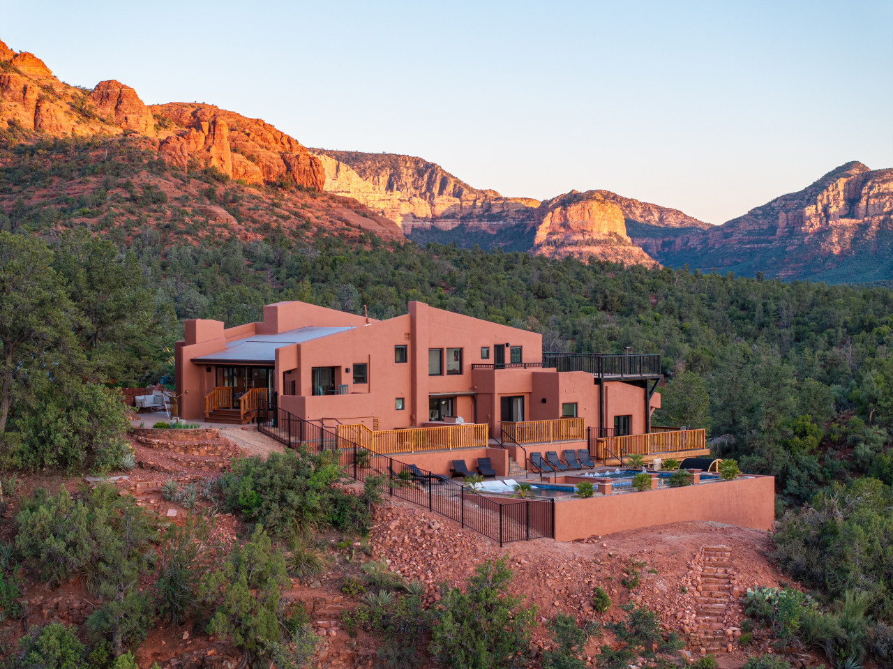 Modern multi-level Sedona rental home surrounded by lush forest and iconic red rock formations at sunset.