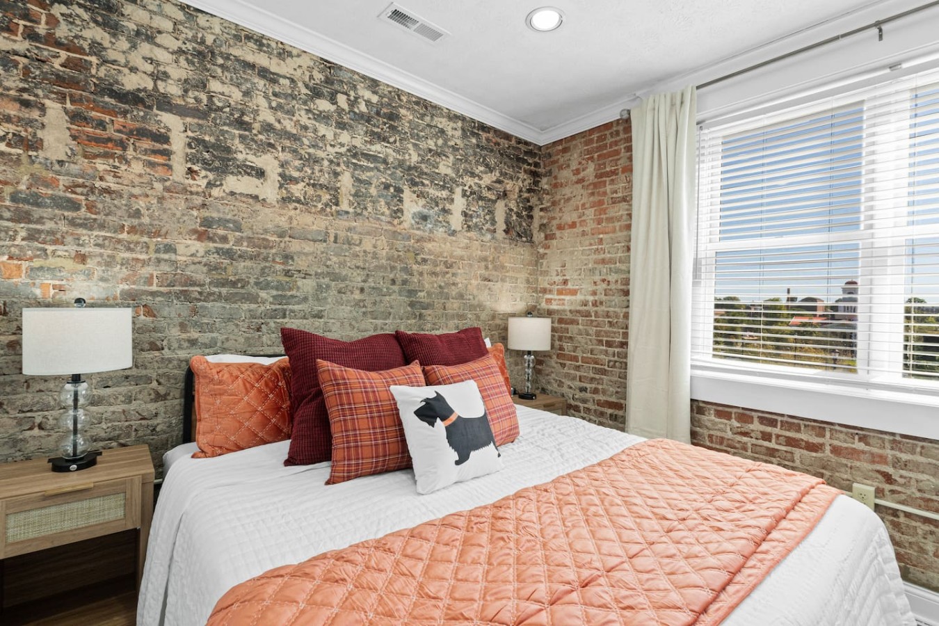 Wake up to natural light streaming through large windows in this cozy bedroom. The king-size bed, accented by warm-toned pillows and an exposed brick wall, offers a relaxing, rustic atmosphere that blends comfort with historic charm.