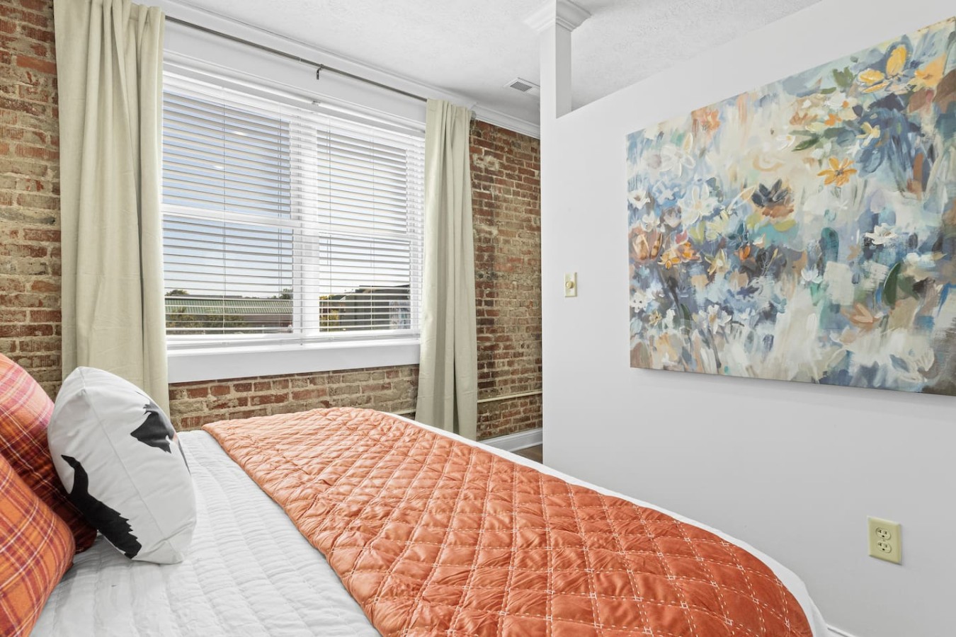Relax in this cozy bedroom, featuring an inviting king-size bed with colorful bedding and soft pillows. The large window fills the space with natural light, while the exposed brick wall and vibrant artwork create a warm and welcoming atmosphere.