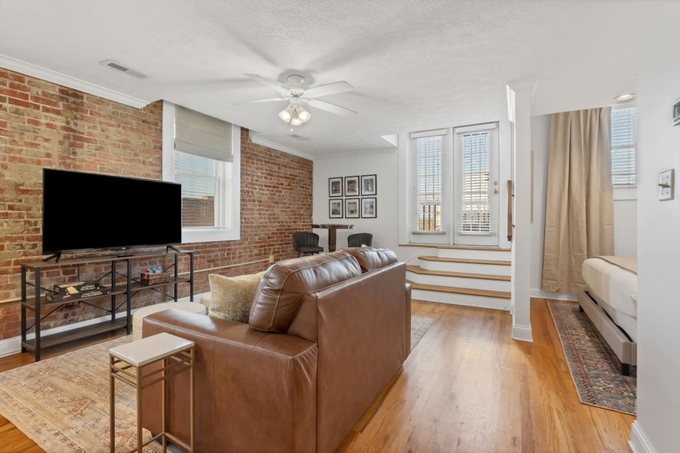Spacious living area with exposed brick, a plush leather sofa, and a large TV. Natural light fills the open layout, featuring a cozy nook and steps to the bedroom. A stylish blend of comfort and charm!