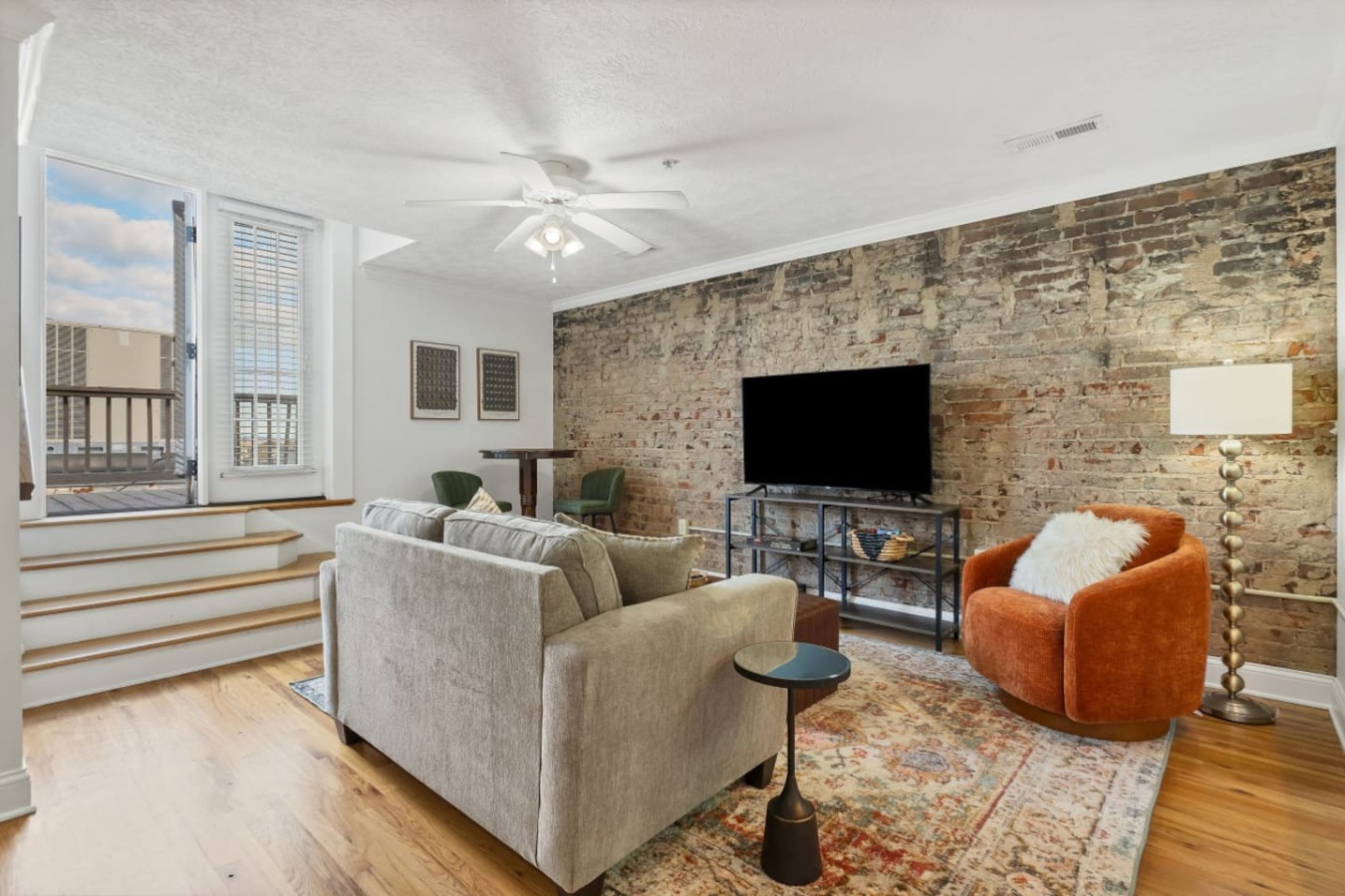 Bright and inviting living room with exposed brick, cozy seating, and a large TV. French doors open to a private balcony, bringing in natural light. Perfect for relaxing after exploring downtown Maryville's shops, restaurants, and local attractions.