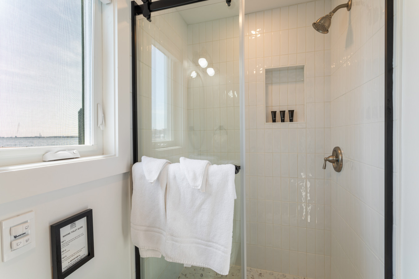 Spacious walk-in shower with a built-in niche for toiletries, barn-door glass, and sunlight streaming in from the harbor.