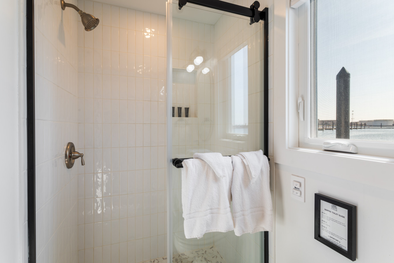 Even the shower has a view — barn-door style glass enclosure, white tile, and natural light from the harbor window.
