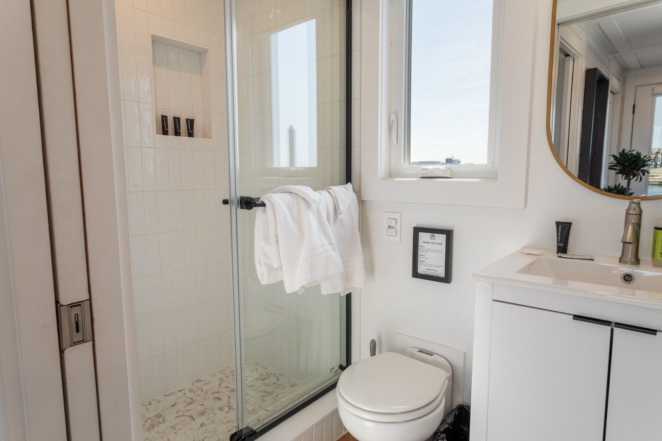 Clean and modern bathroom with walk-in glass shower, designer vanity, gold mirror, and even a water view from the window.