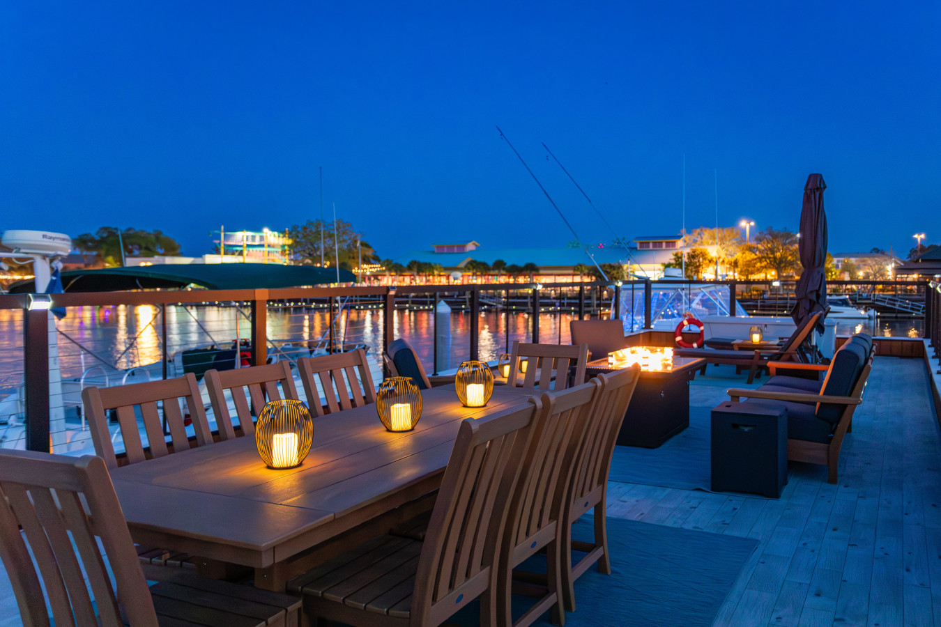 Rooftop dining table at blue hour with lanterns glowing, the fire pit lit in the background, and the Barefoot Landing waterfront string lights reflecting across the Intracoastal after dark.