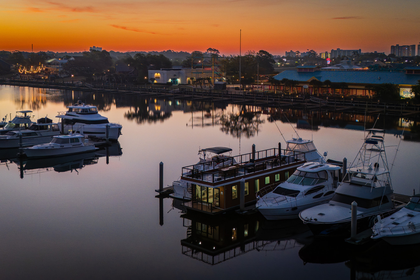 Drone view of Palmera at sunrise with the Intracoastal Waterway perfectly still, the Barefoot Landing shops and string lights reflected on the water, and the Grand Strand skyline glowing along the horizon.