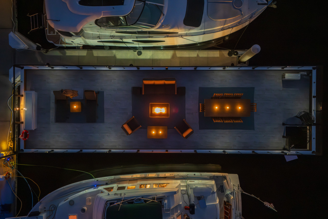 Overhead drone view of the rooftop at night showing the full layout from above: fire pit glowing at center, lounge seating, dining table, and chaise lounges, flanked by boats docked on either side.