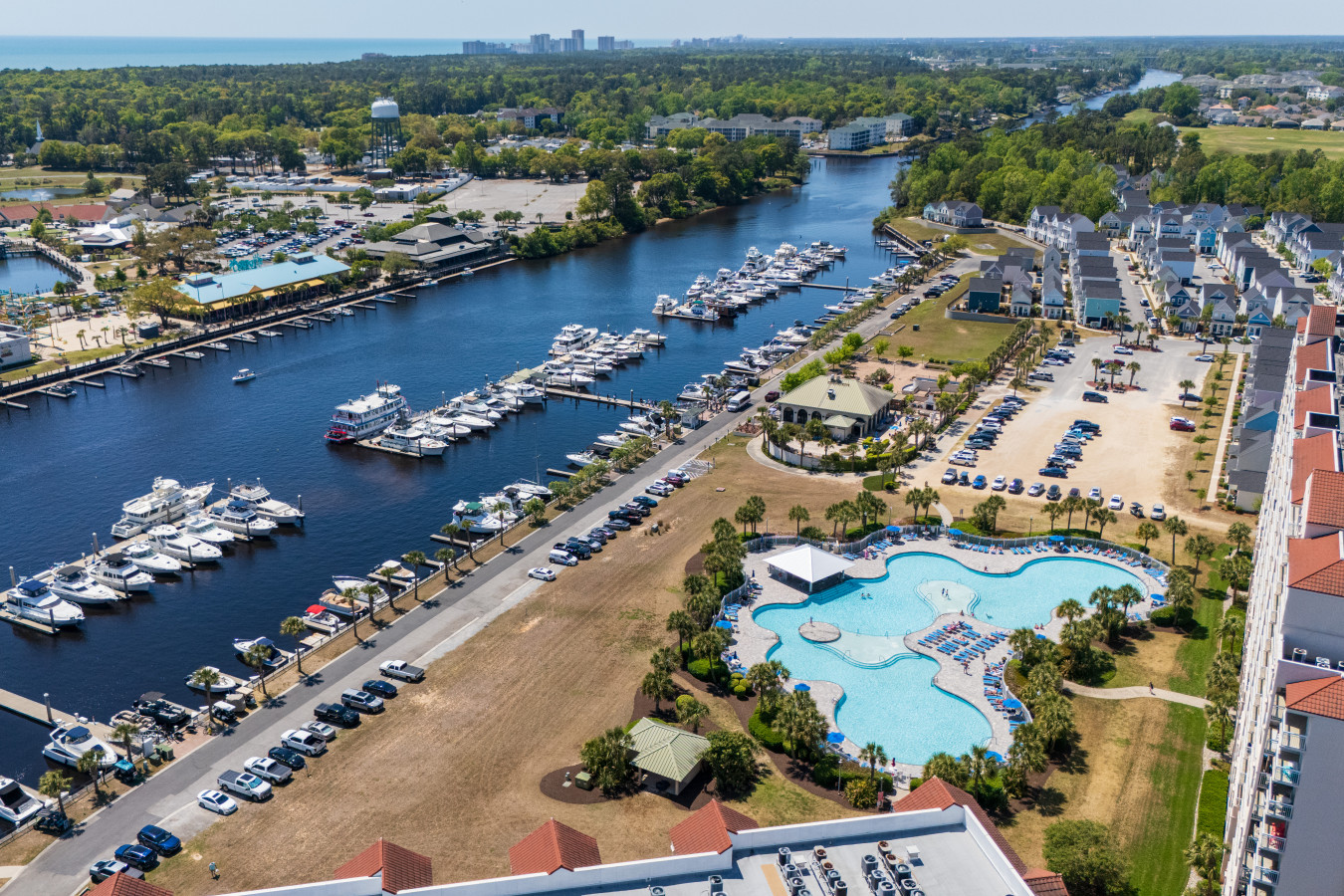 Aerial view of Barefoot Landing Marina, the Intracoastal Waterway, and the resort pool included with your stay, with the Atlantic Ocean and Grand Strand skyline visible in the distance.