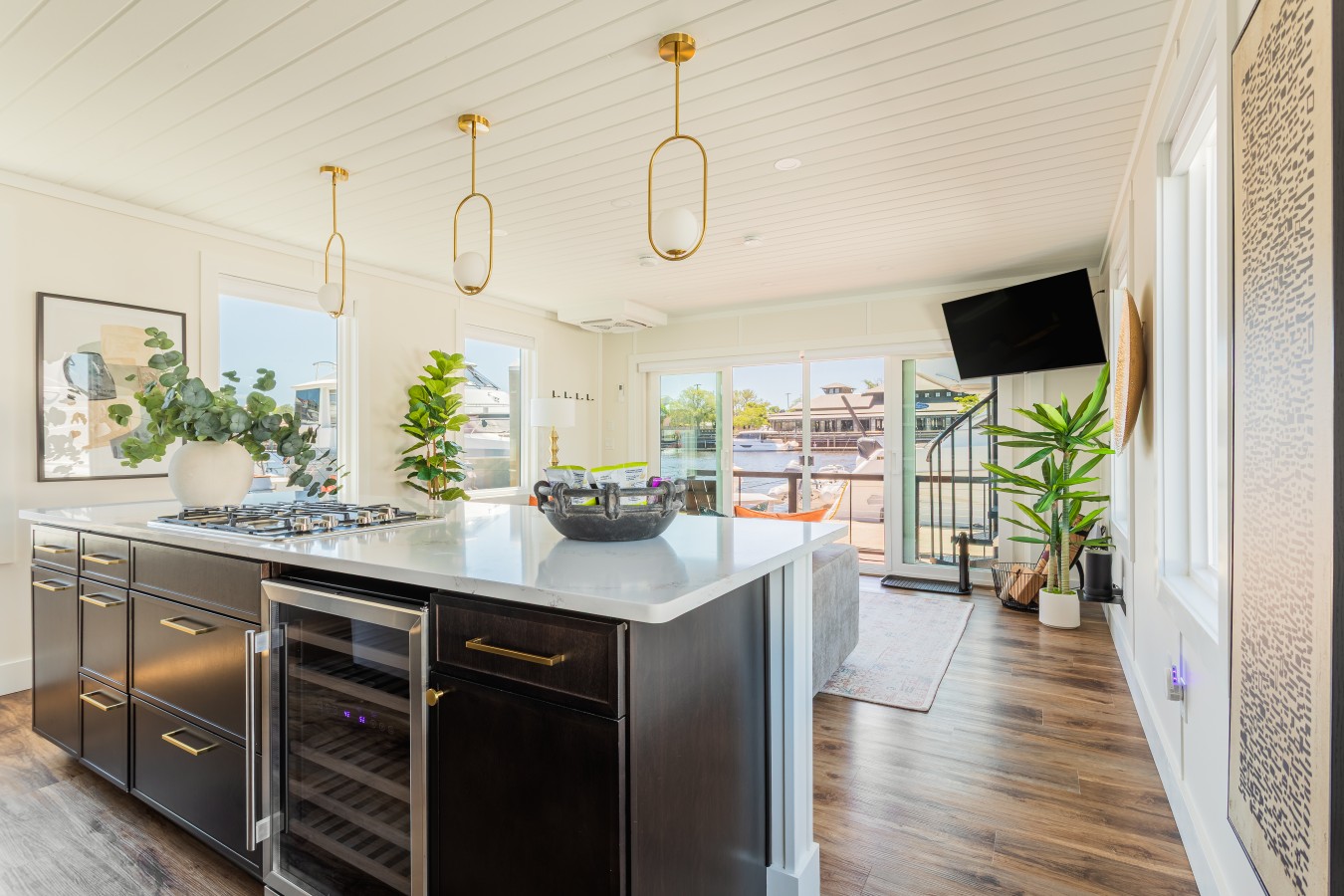 The kitchen island with quartz counters, a gas cooktop, wine fridge, brass hardware, and gold pendant lighting, opening directly into the living area with marina views through the sliding glass doors.