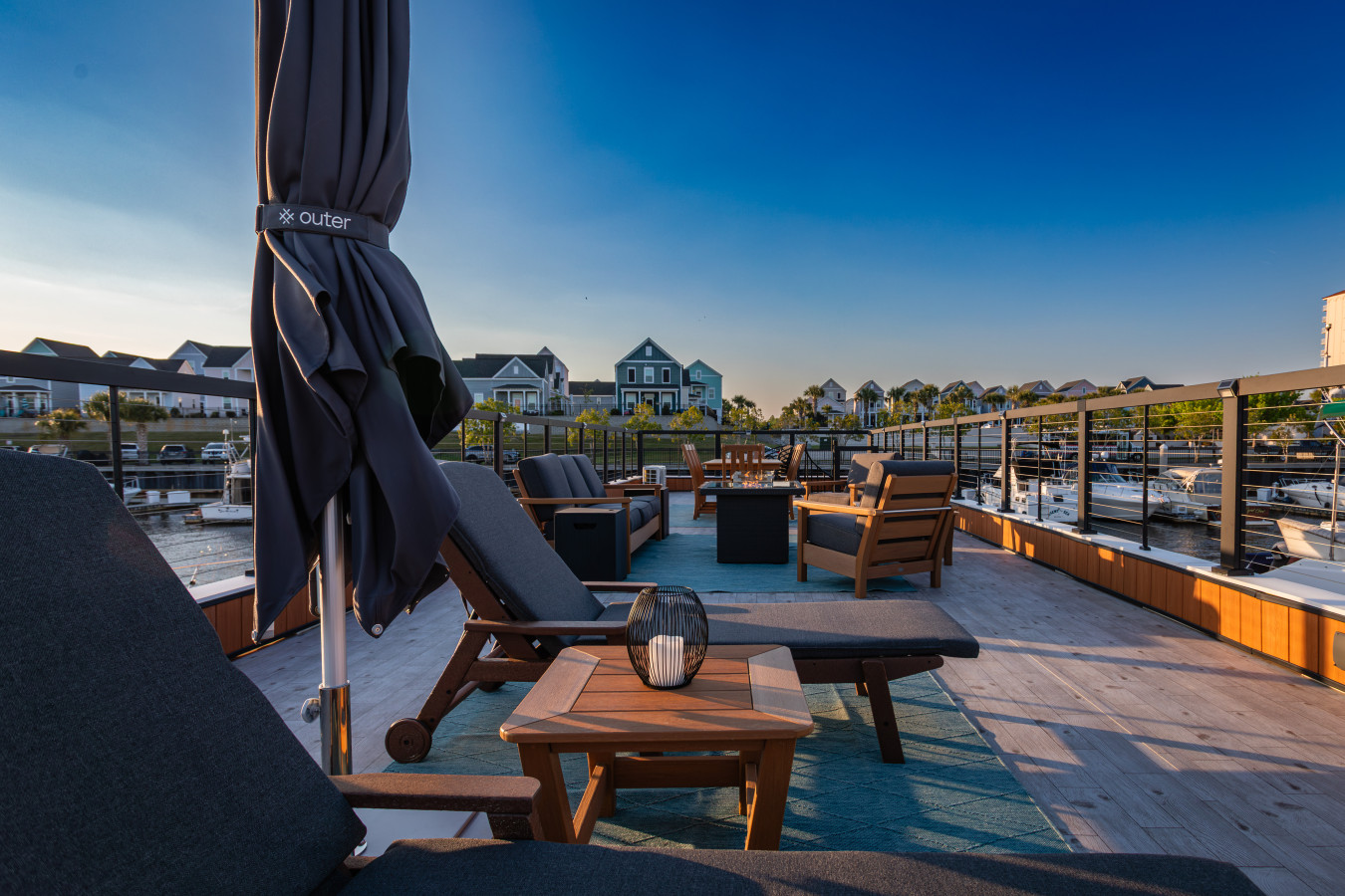 Rooftop chaise lounges with umbrella, fire pit, and deep seating looking across the marina toward the colorful waterfront homes of Barefoot Landing under a clear coastal sky.