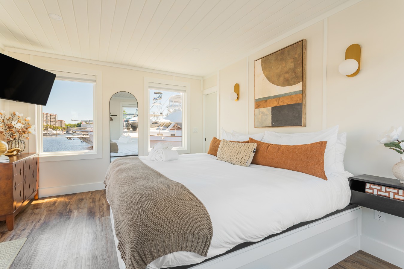 The king bedroom with marina views on two sides, warm terracotta linens, gold sconce lighting, an arched mirror, and contemporary artwork above the bed.