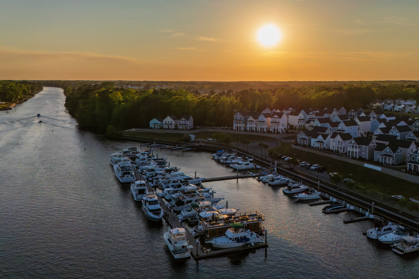Drone view of Palmera at sunset from above the Intracoastal Waterway, the rooftop visible among the boats, with the coastal homes of Barefoot Landing and a golden sky stretching over the tree line.