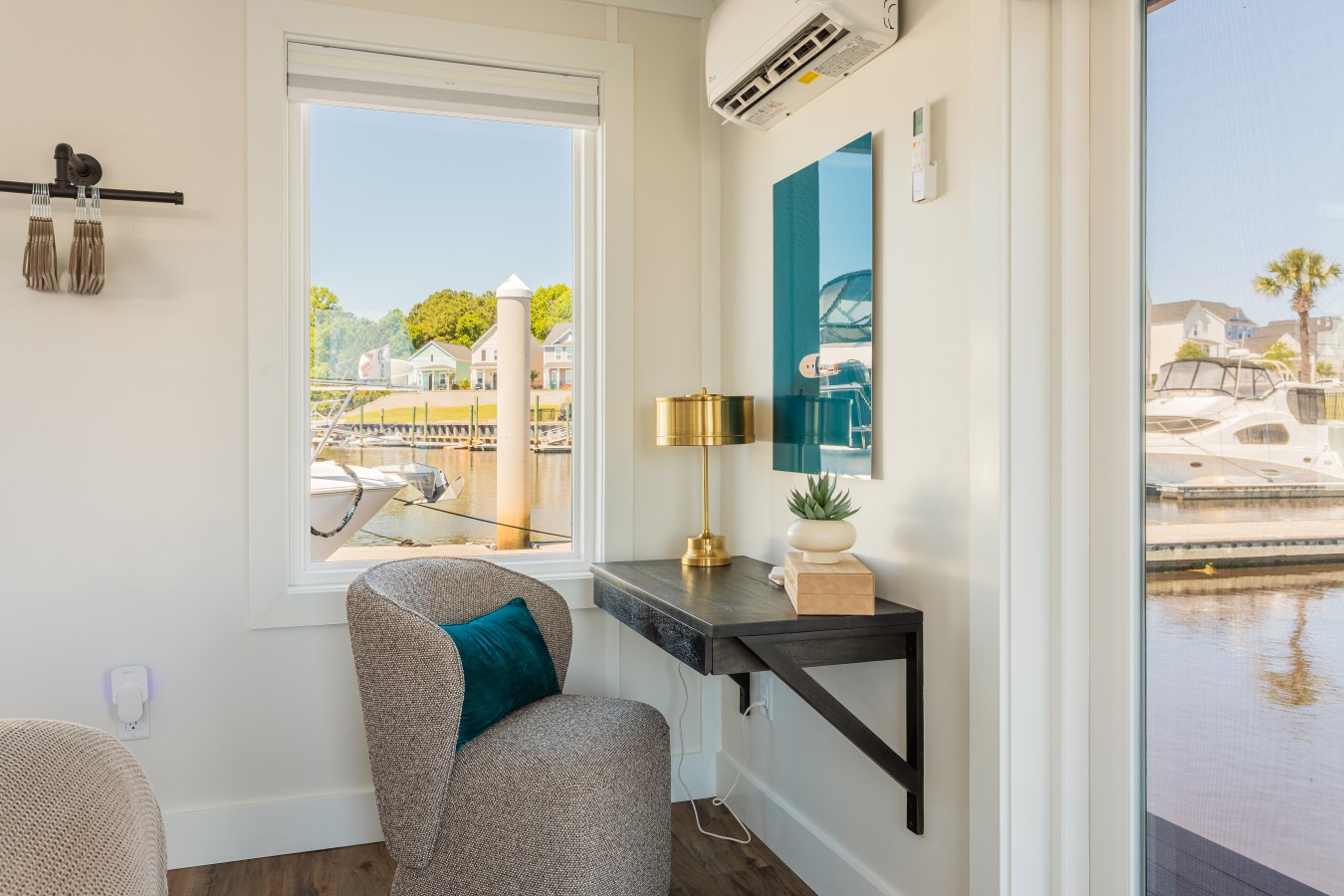The bedroom workspace corner with a floating desk, brass lamp, lounge chair, teal accent pillow, and marina views through both the window and the floor-to-ceiling glass door.