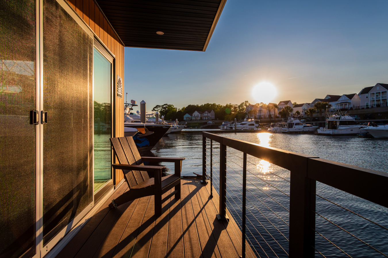 Front deck at sunset with an Adirondack chair, the FLOHOM logo on the glass door, and the sun dropping over the Intracoastal Waterway as light catches the water and the marina beyond.