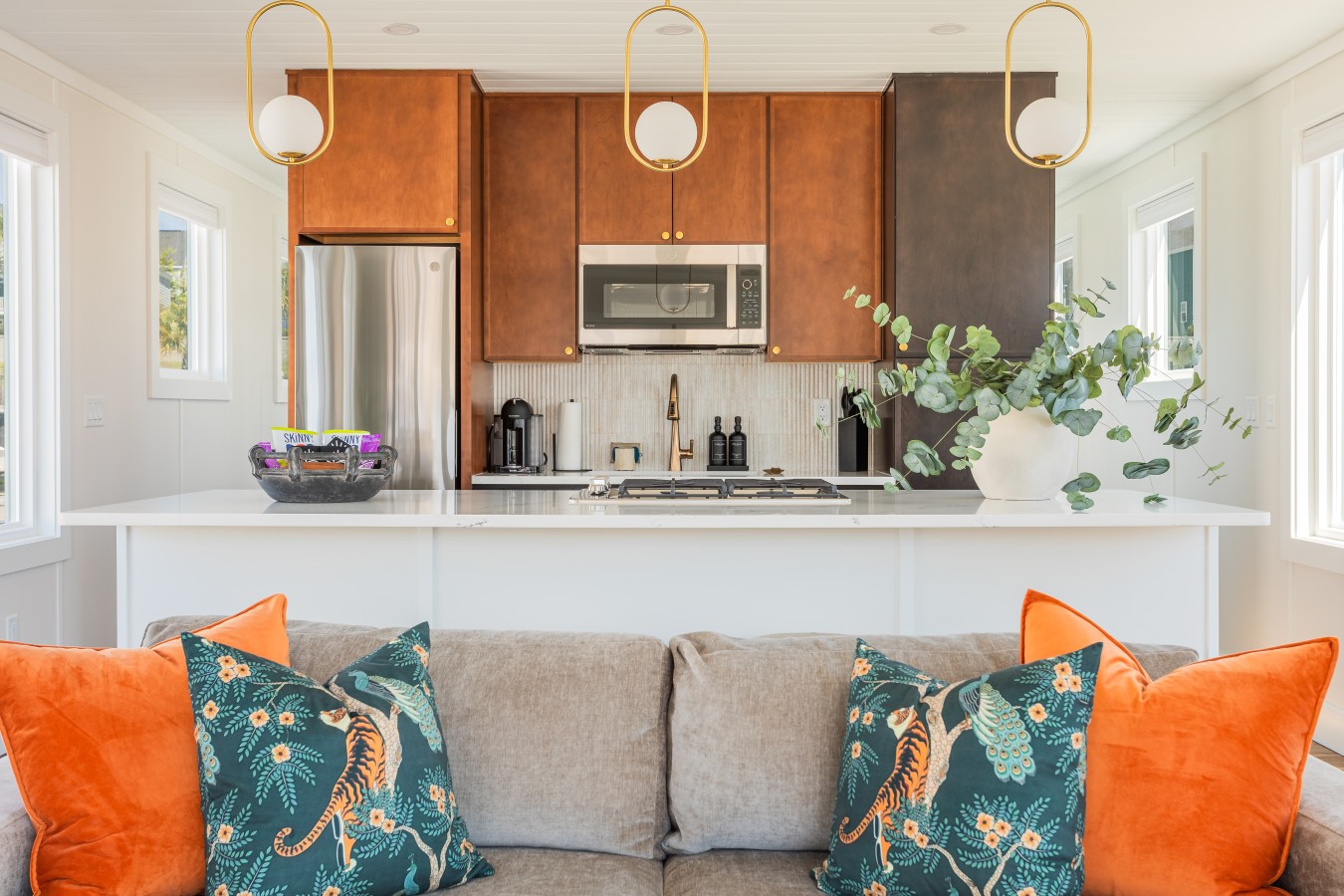 The kitchen from the living area, with warm cedar cabinetry, gold pendant lighting, a quartz island, brass faucet, and bold botanical pillows anchoring the sofa in the foreground.