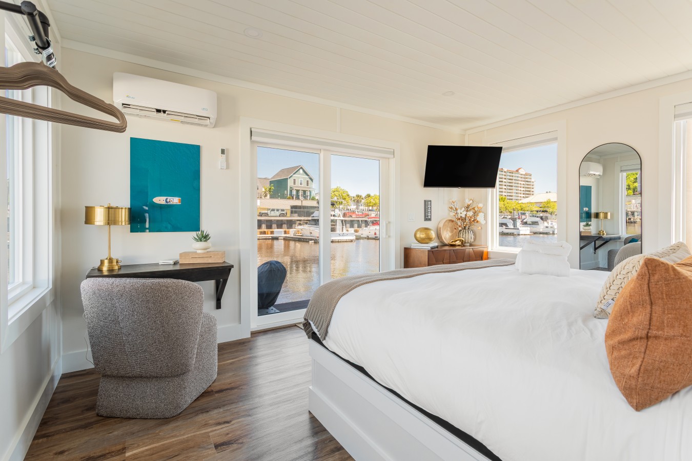 The king bedroom looking toward the sliding glass door, with direct marina views on three sides, a dedicated workspace, lounge chair, wall-mounted TV, and the Barefoot Resort visible across the waterway.