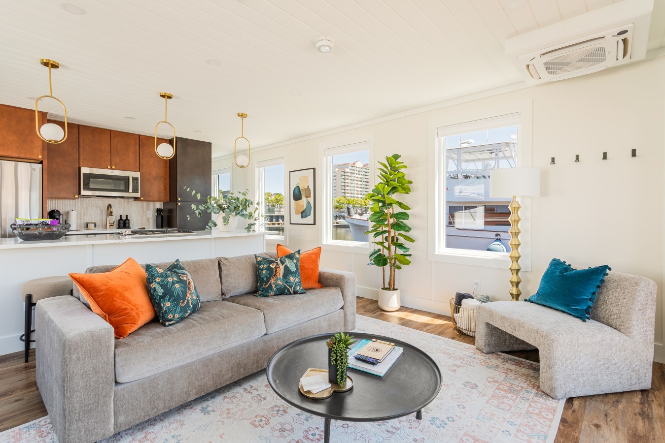 The living area from the entry side, with the sofa, lounge chair, bold botanical pillows, marina-view windows, the Barefoot Resort visible across the water, and the cedar kitchen with gold pendants beyond.