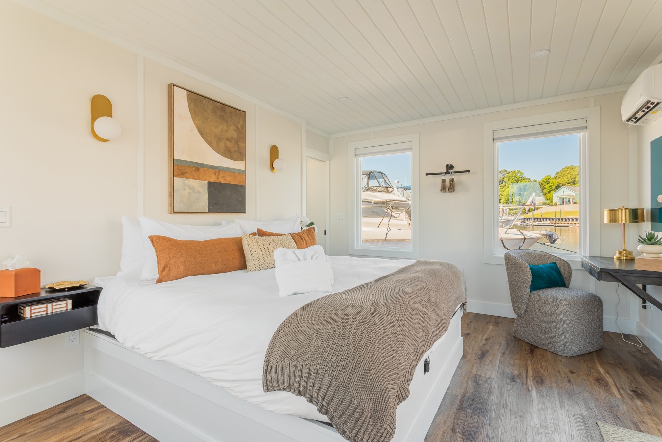 The king bedroom from the foot of the bed, with marina views through both windows, gold sconce lighting, contemporary artwork, a dedicated workspace, and a lounge chair in the corner.