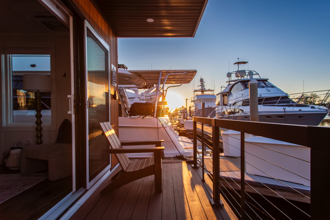 Palmera's entry deck at sunrise, with warm cedar finishes, an Adirondack chair, cable railing, and the sun rising down the dock between the yachts of Barefoot Marina.