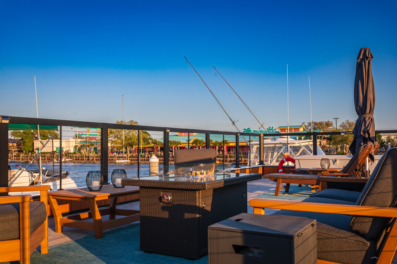 Fire pit close-up on the rooftop with the flame lit, lounge seating on both sides, and the Barefoot Landing waterfront shops and restaurants visible directly across the Intracoastal.