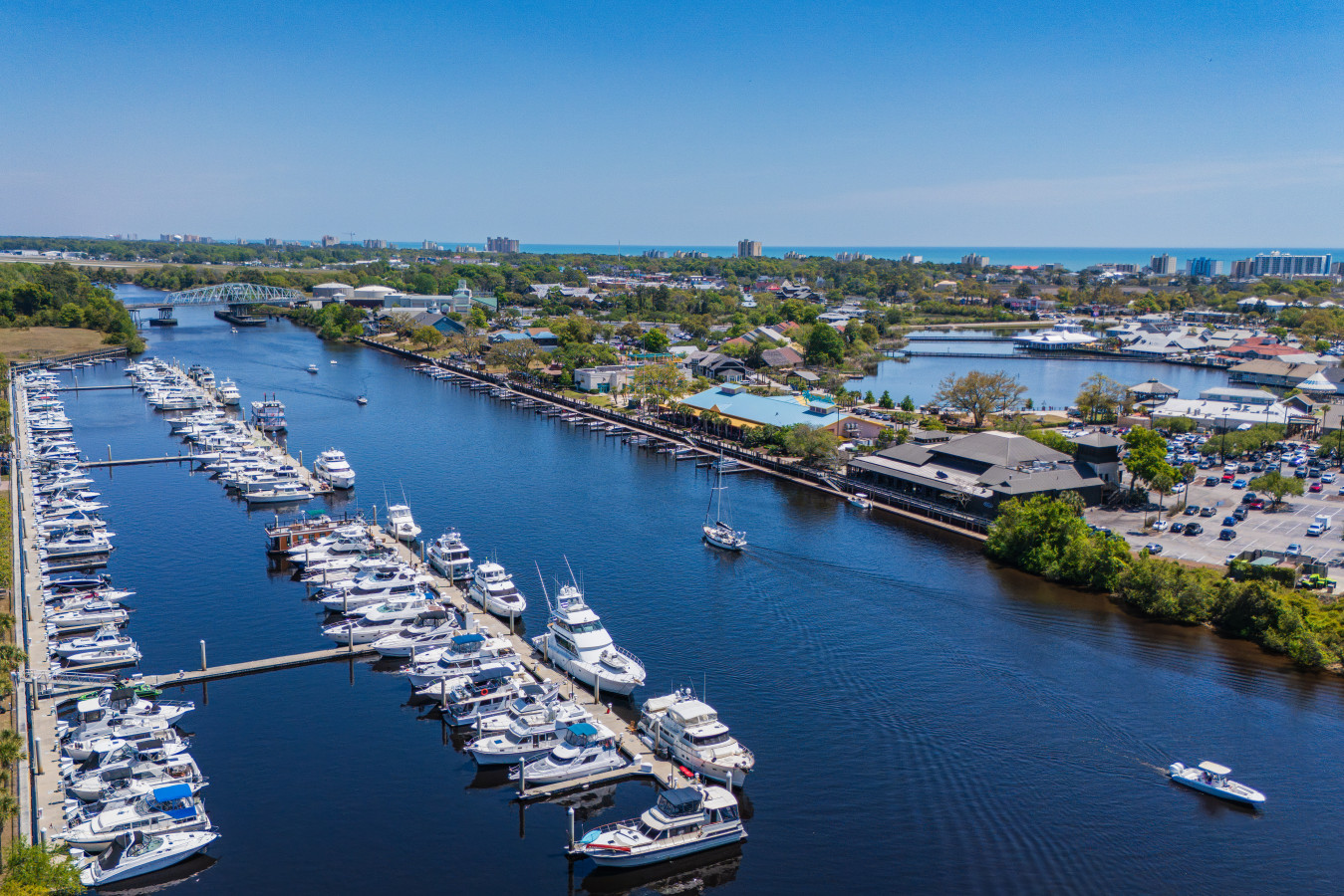 Wide aerial view of Barefoot Landing Marina and the Intracoastal Waterway looking toward the Atlantic Ocean, with Barefoot Landing shops and restaurants lining the waterfront and boats cruising the channel.