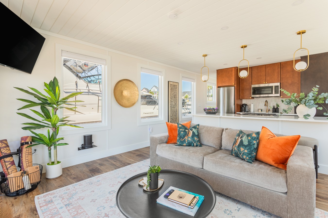 The open living area looking toward the kitchen, with bold botanical pillows, marina-view windows, gold pendant lighting over the cedar cabinetry, and the quartz island anchoring the space beyond.