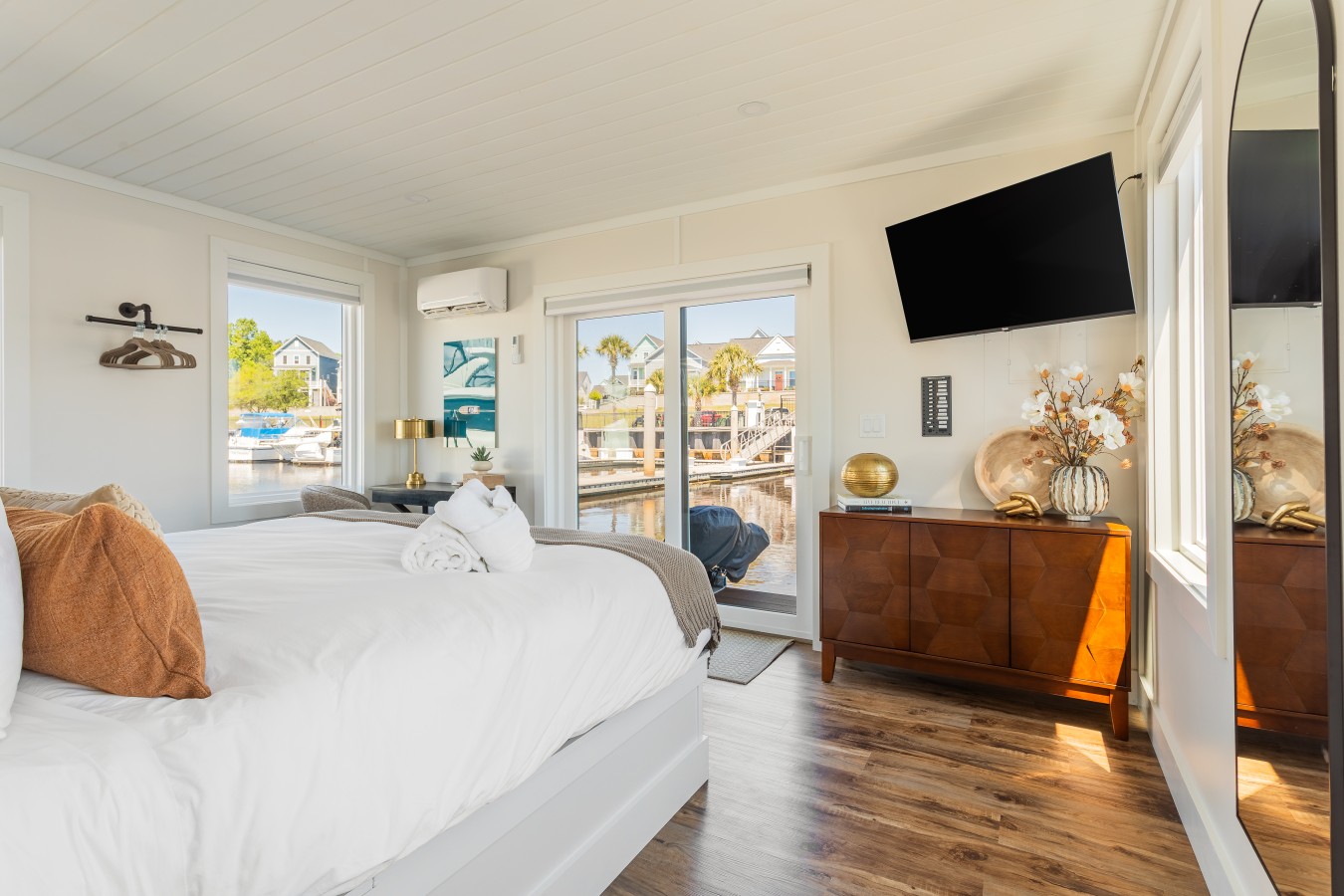 The king bedroom with a sliding glass door opening directly to the marina, a geometric walnut dresser, wall-mounted TV, and the colorful waterfront homes of Barefoot Landing visible across the Intracoastal.